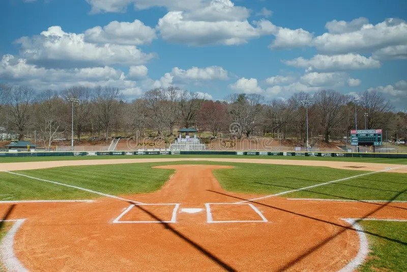 View of a baseball field from home plate with pitcher’s mound and outfield under a bright blue sky with clouds.